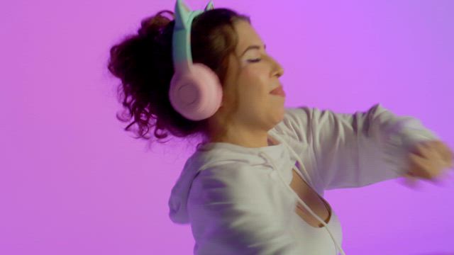 A young smiling woman wearing flickering headphones with kitty ears is dancing energetically over a colorful purple background.