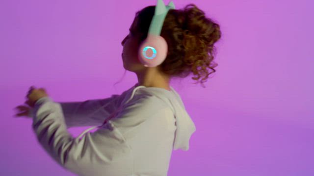 A young woman wearing white clothes and flickering RGB headphones joyfully dances over a purple background.