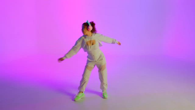 A young woman wearing all white clothes and flickering RGB headphones dances gracefully over a vibrant purple hue backdrop.