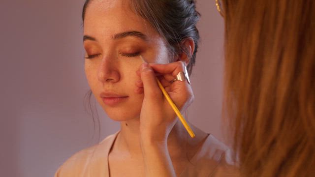 A skilled makeup artist add final touches to the eyelashes of a young serene model in a softly lit studio.