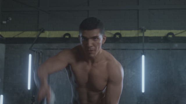 A young shirtless muscular man exercises moving the battle ropes with determination at the gym.