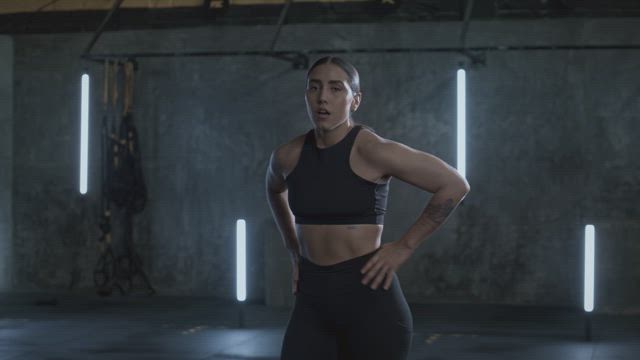 At a pause on her fitness training a young muscular woman wearing a snugly sportsware black outfit is inhaling and exhaling at the gym.