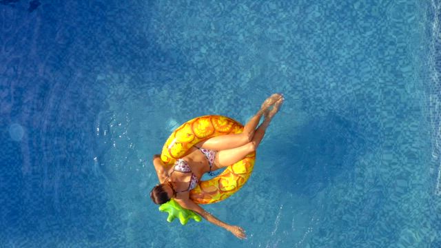 A young woman seen from above on a pineapple floaty enjoys the swimming pool on a sunny day.