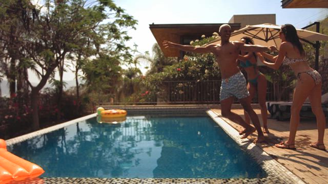 In slow motion two young woman wearing beach bikinis push a young African American man towards the pool creating a refreshing splash on a sunny summer day.