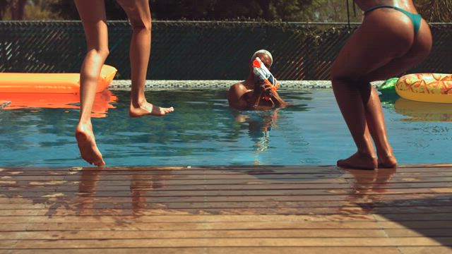 On a display of leisure and fun summer vibes a young man aims with a water gun at two young girls wearing bikinis jumping at the pool.